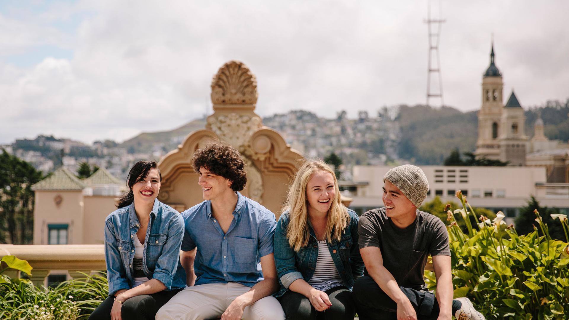 Four students sit on a bench with a view of campus in the background