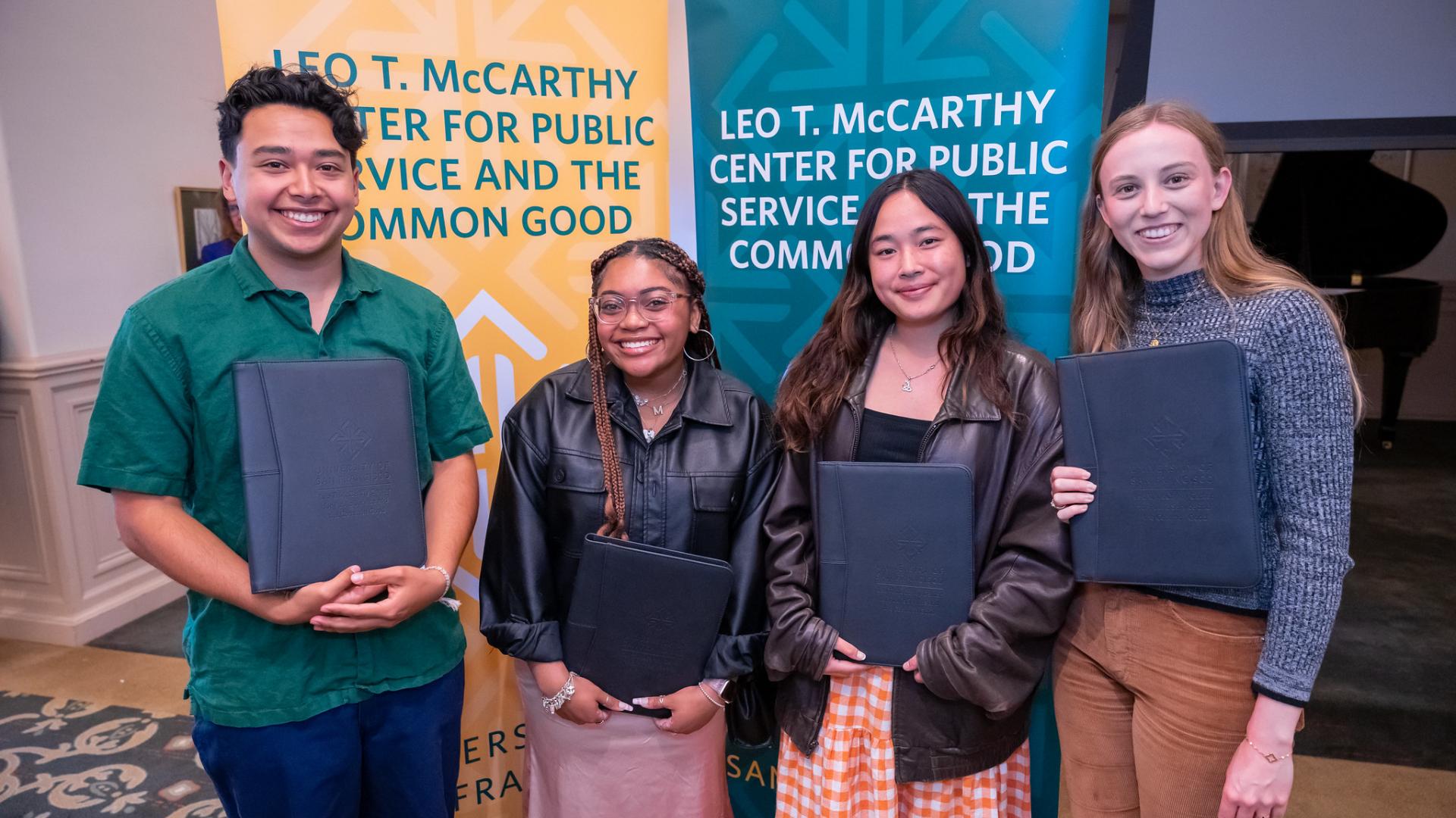 four students holding padfolios in front of two banner signs indicating Leo T McCarthy Center