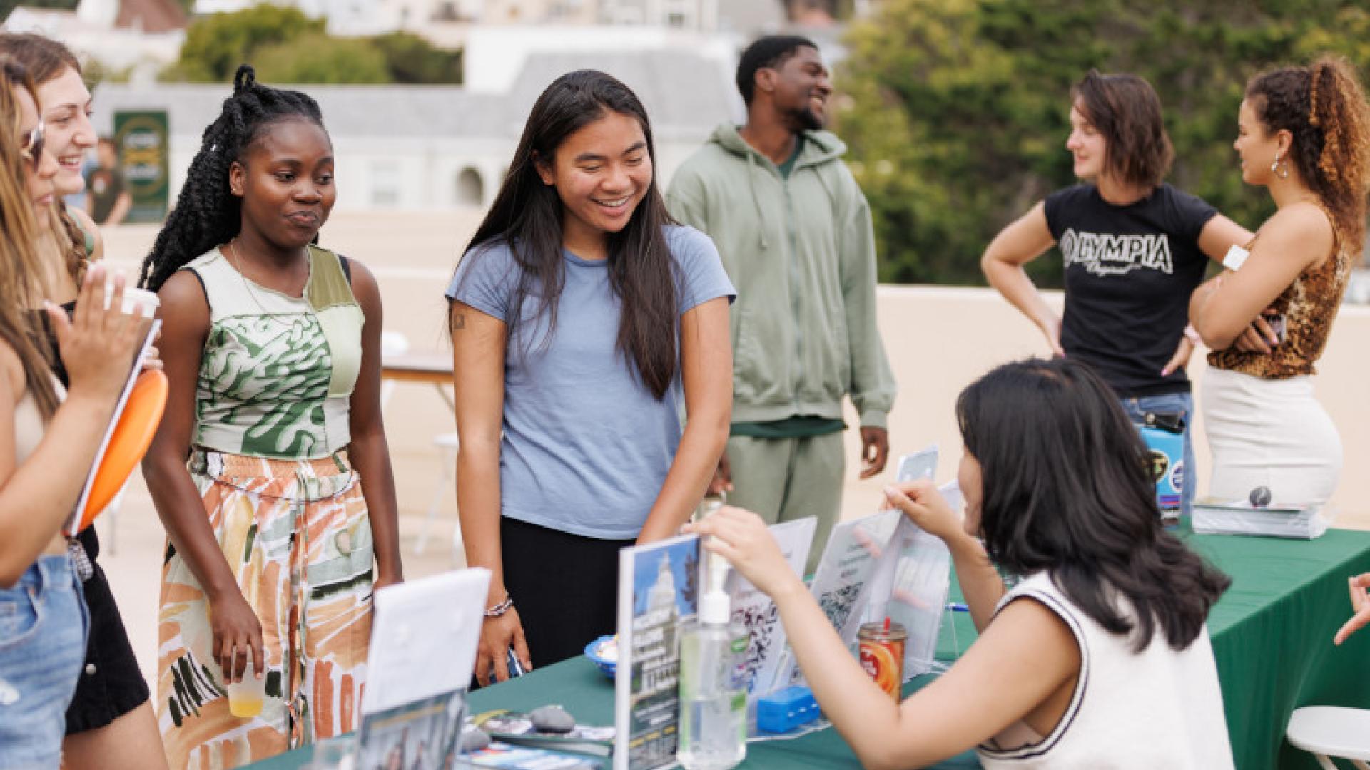 4 students standing and listening to representative seated at a tabling event.