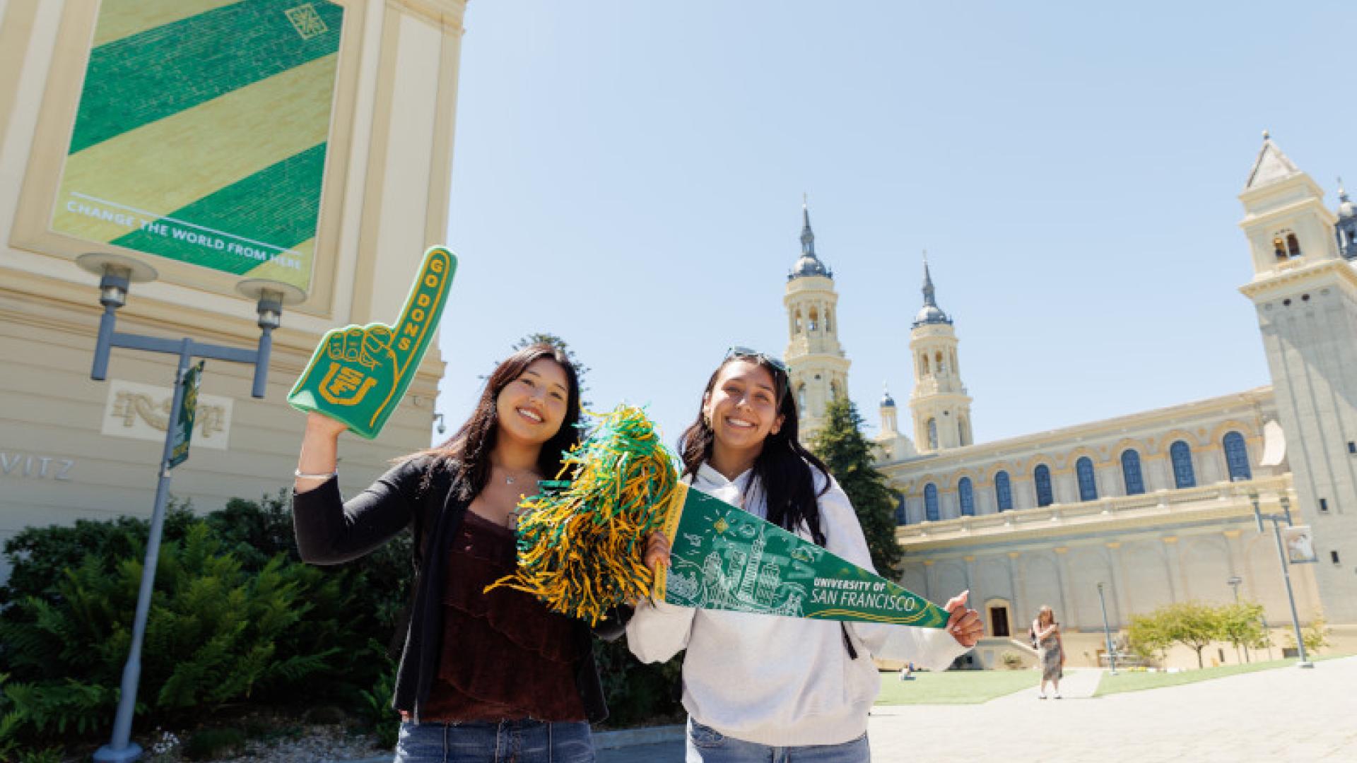 Two students holding a USF foam finger, a gold and green pom-pom and a USF pendant.
