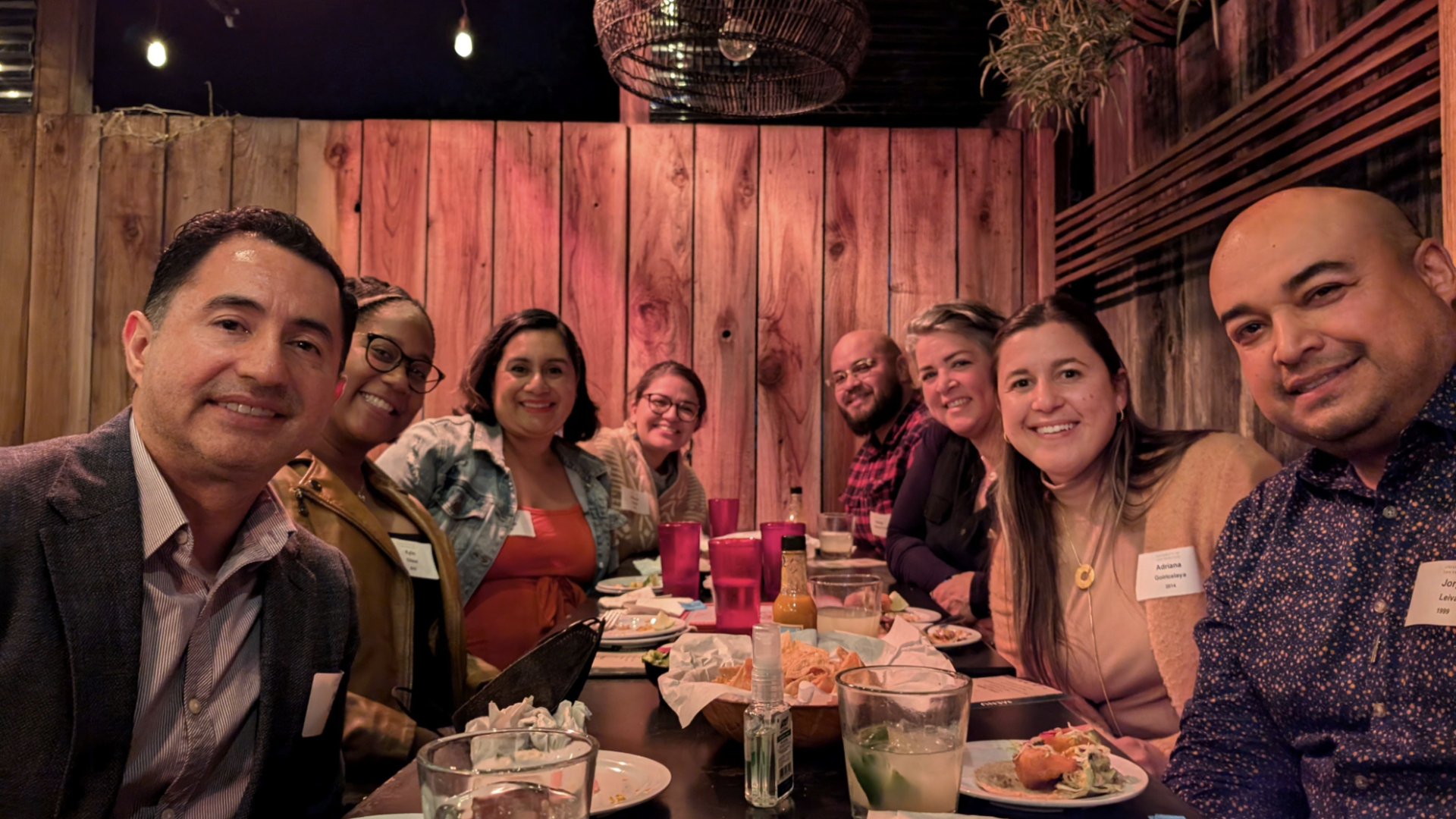 Group of 8 gathered around a table eating and posing for photo