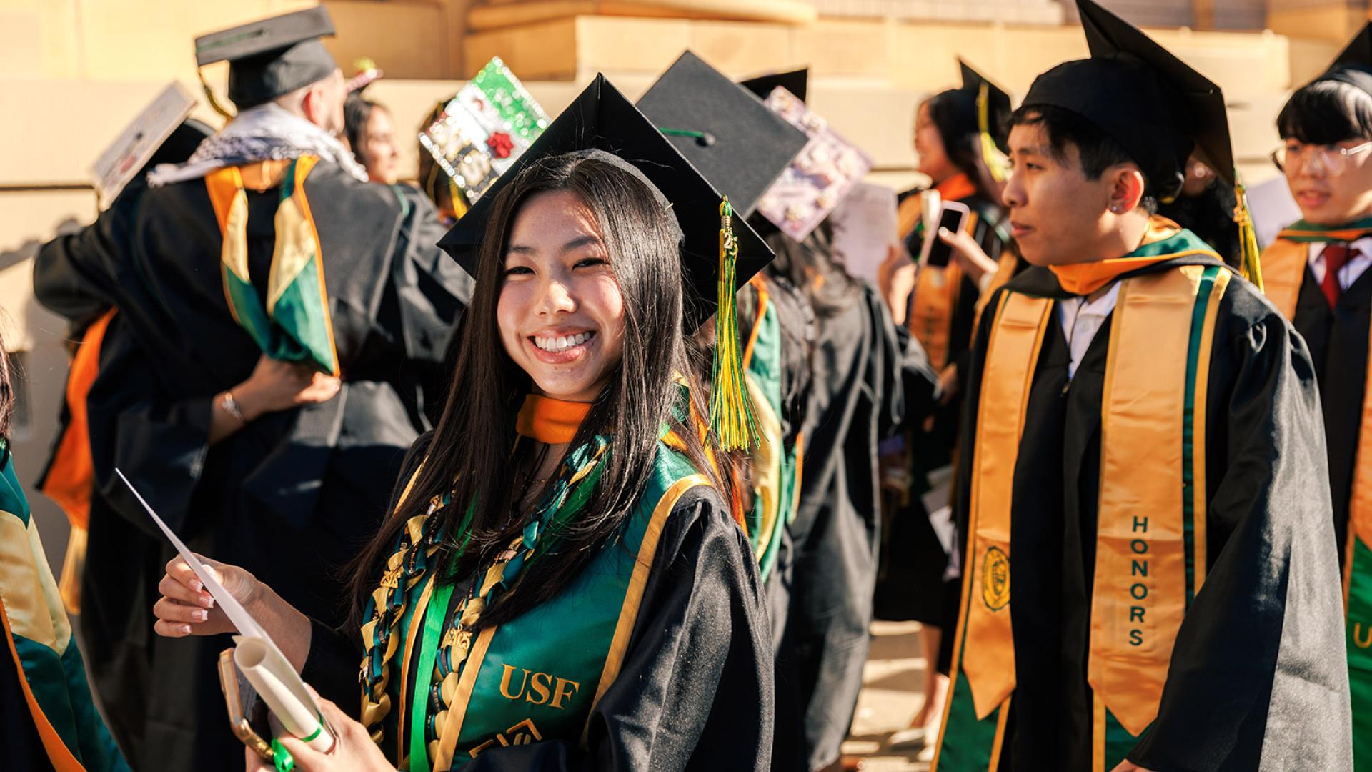 student in gap in gown stands among a crowd of other graduates outside on usf campus 
