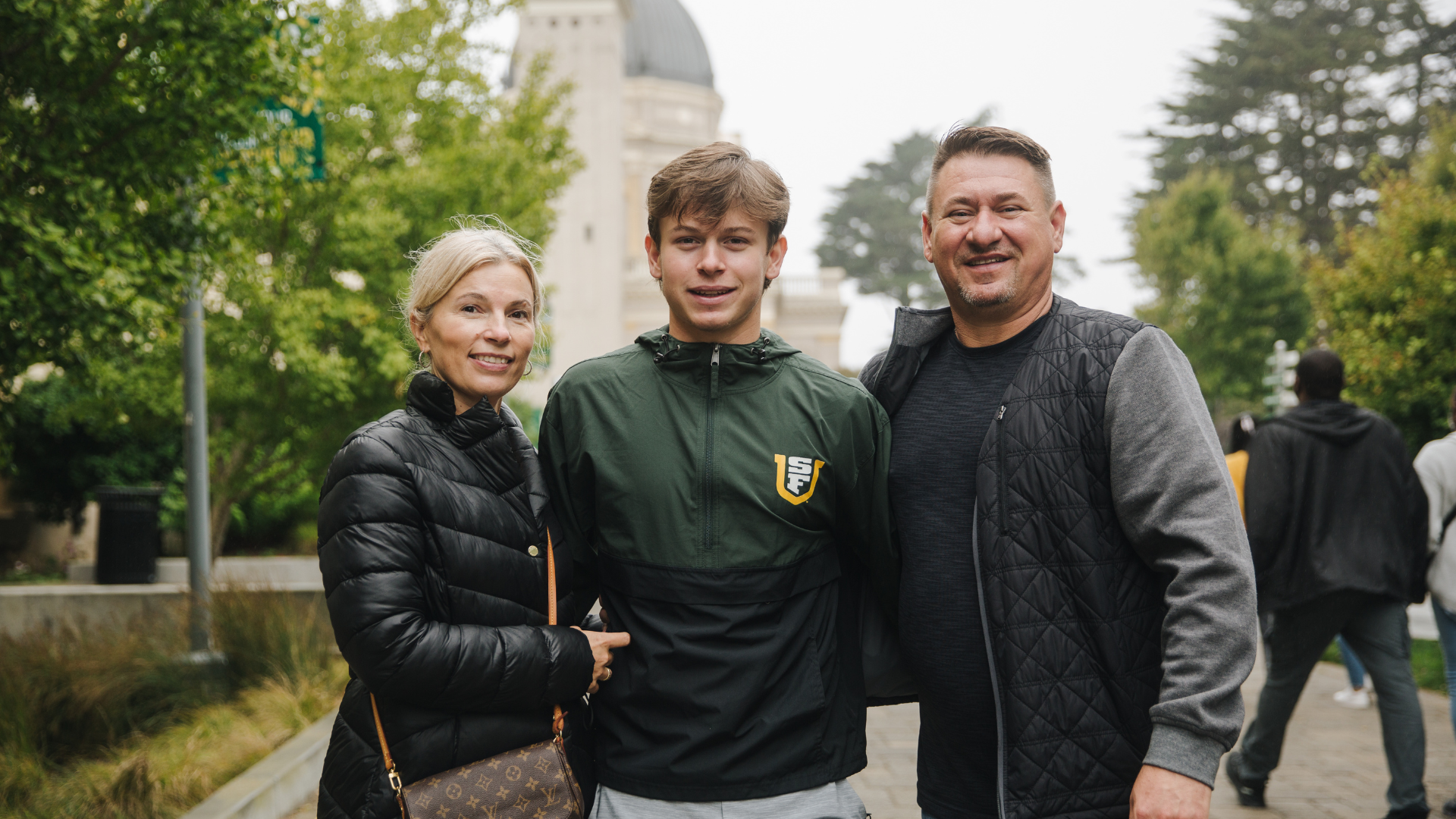 A student poses with two family members outdoors on the University of San Francisco campus, wearing a green USF jacket as the group smiles for the camera.