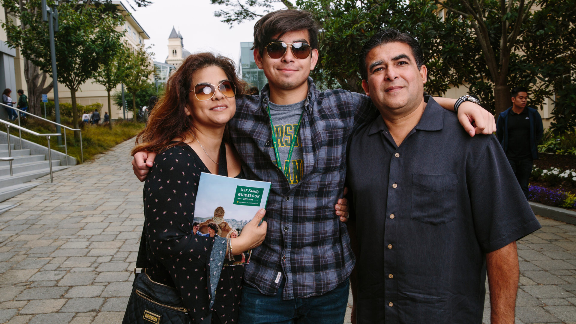 A student stands with two family members outdoors on the University of San Francisco campus, with his arms around them as they pose together. One family member holds a USF Family Guidebook.