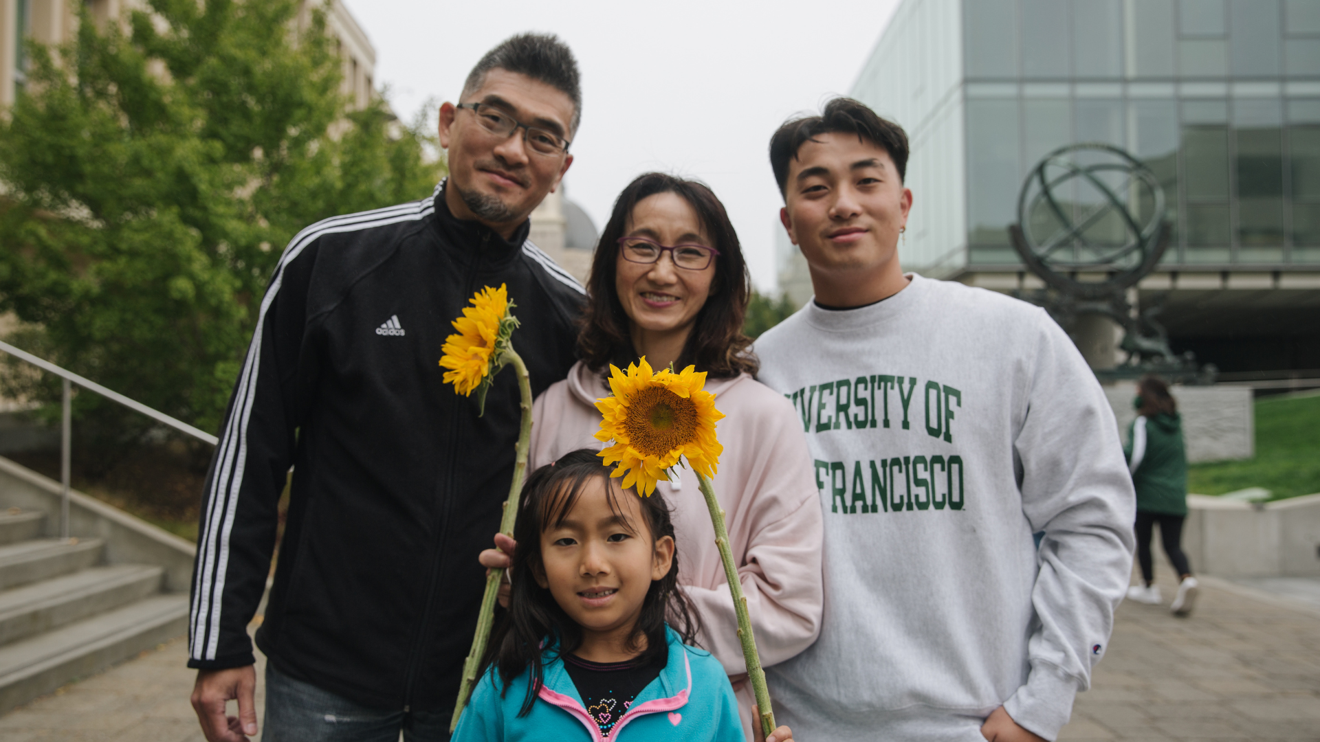 A family of four poses together outdoors on the University of San Francisco campus. Two family members hold bright yellow sunflowers, and one person wears a University of San Francisco sweatshirt.