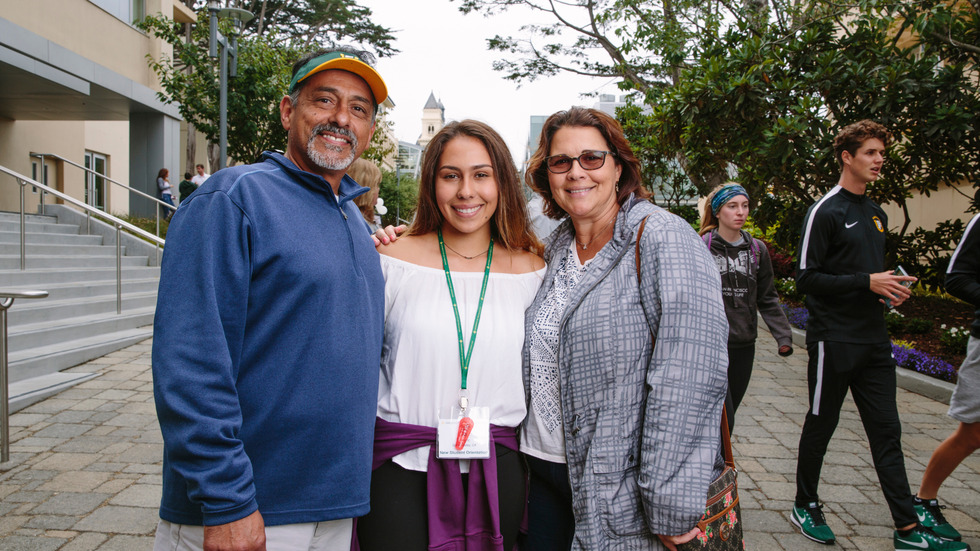 A student stands between two family members as they smile for a photo outdoors on the University of San Francisco campus during New Student Orientation.