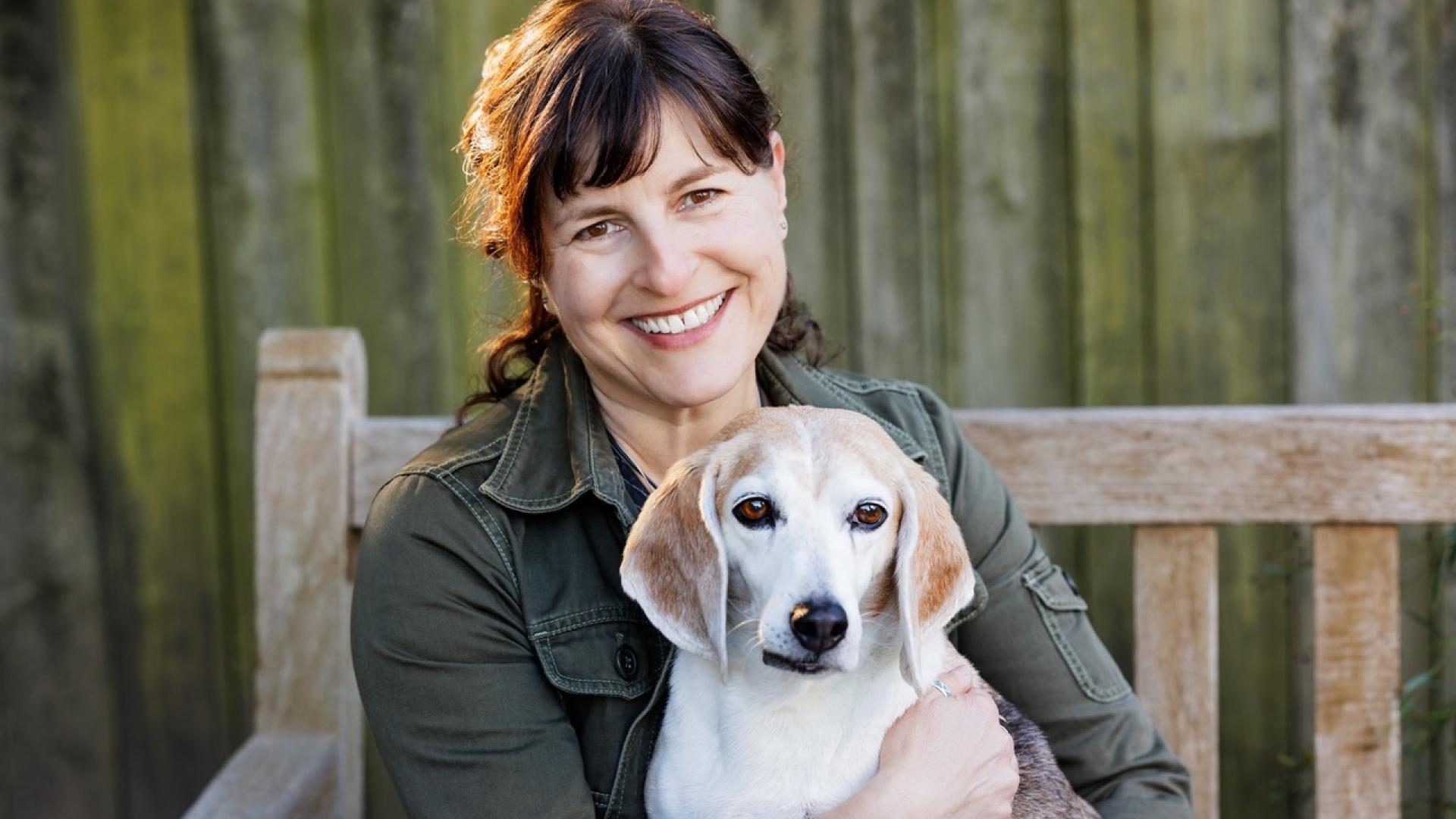A smiling woman with dark brown hair and bangs, wearing an olive green utility jacket, sits on a weathered wooden bench and holds a white and tan beagle in her lap. The dog looks toward the camera with soft brown eyes.